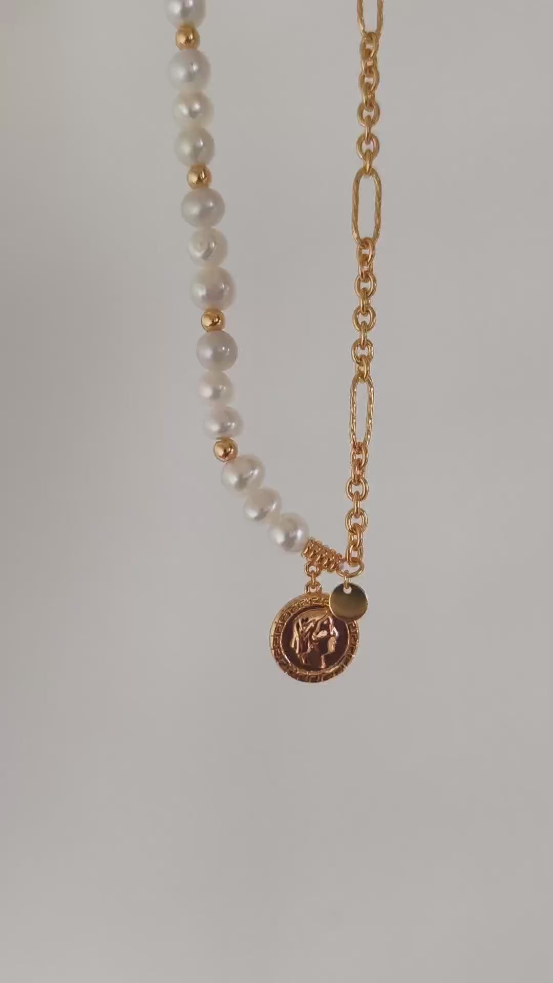 This cinematic close-up video captures the dynamic movement and radiant light of the Calyx asymmetric necklace. The camera pans across the soft, iridescent surfaces of natural baroque pearls, transitioning seamlessly into the high-luster glow of the 18K gold vermeil link chain. The video highlights the intricate relief of the vintage Queen Elizabeth coin pendant, showcasing its artisanal texture as it catches the light. This visual demonstration emphasizes the piece's elegant drape and "half-sweet, half-bol