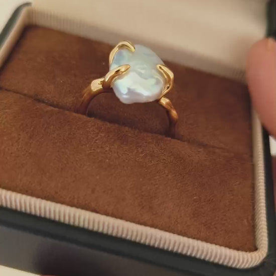 A detailed view of the Aura ring being held, demonstrating the mesmerizing glow of the baroque pearl and the comfort of the adjustable open-band design.