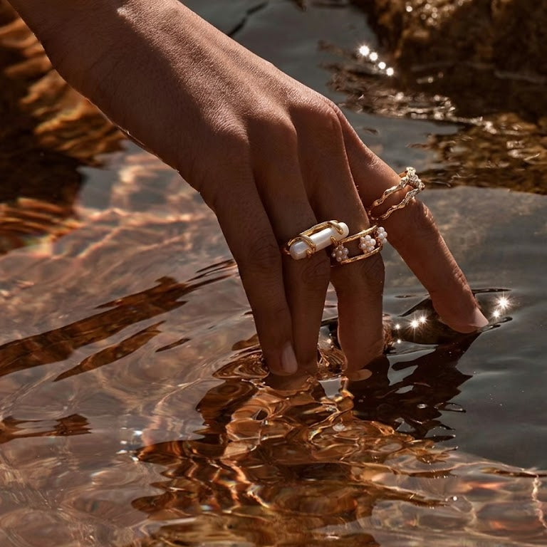Hand wearing gold freshwater pearl rings touching sunlit water, with warm reflections and soft ripples, luxury summer jewelry