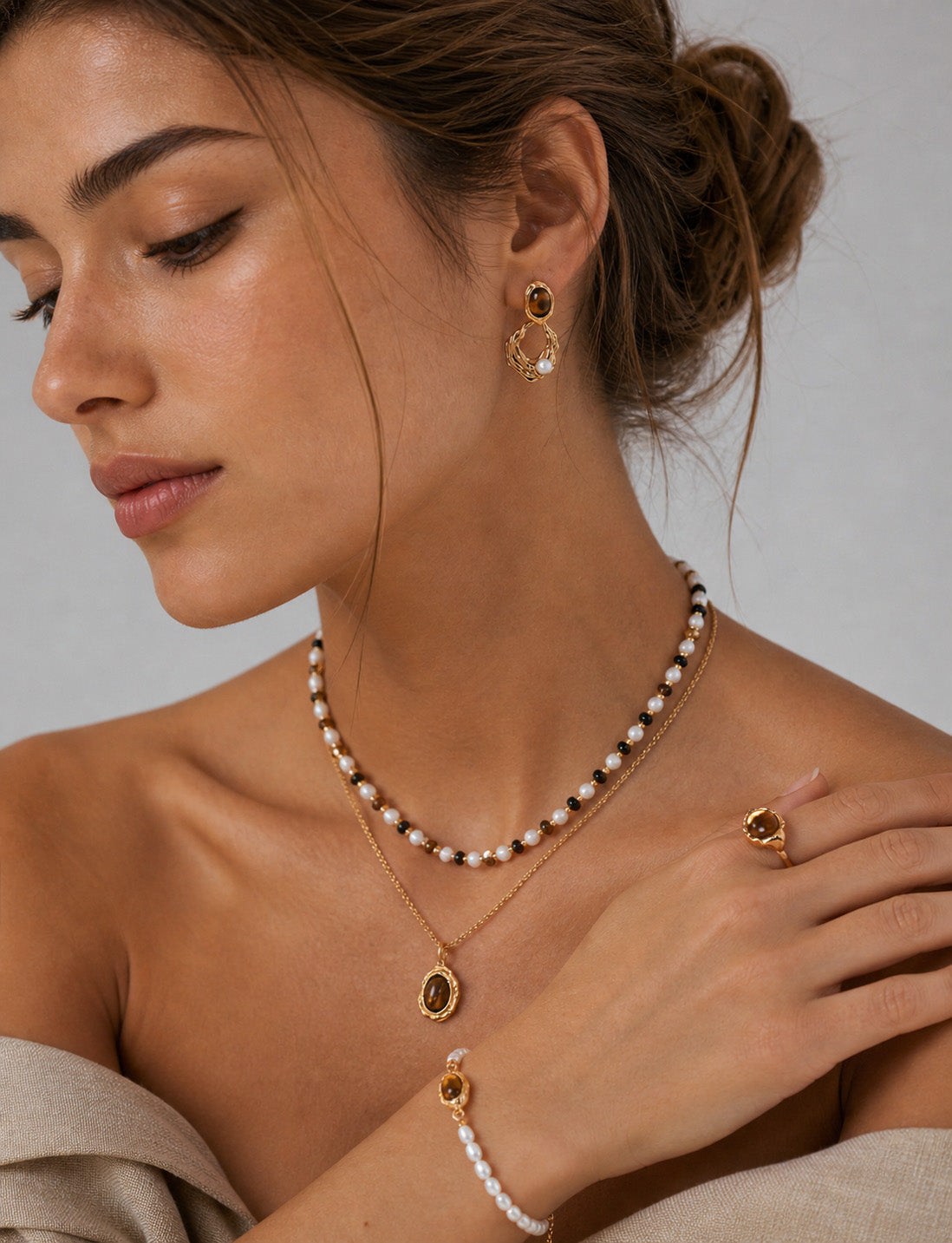A lifestyle shot of the Gaia Tiger's Eye and pearl sets including earrings, rings and necklace styled for a modern bohemian look. The visual demonstrates the 38mm drop length and how the natural stones catch the light to illuminate the face with warm, earthy tones.